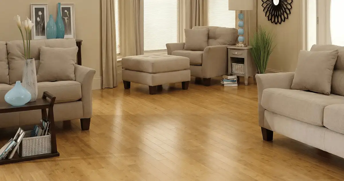 A living room with professionally installed bamboo flooring, beige sofas, a matching ottoman, a side table with a blue lamp, vases, framed pictures, and large windows with blinds and curtains in Aurora, IL